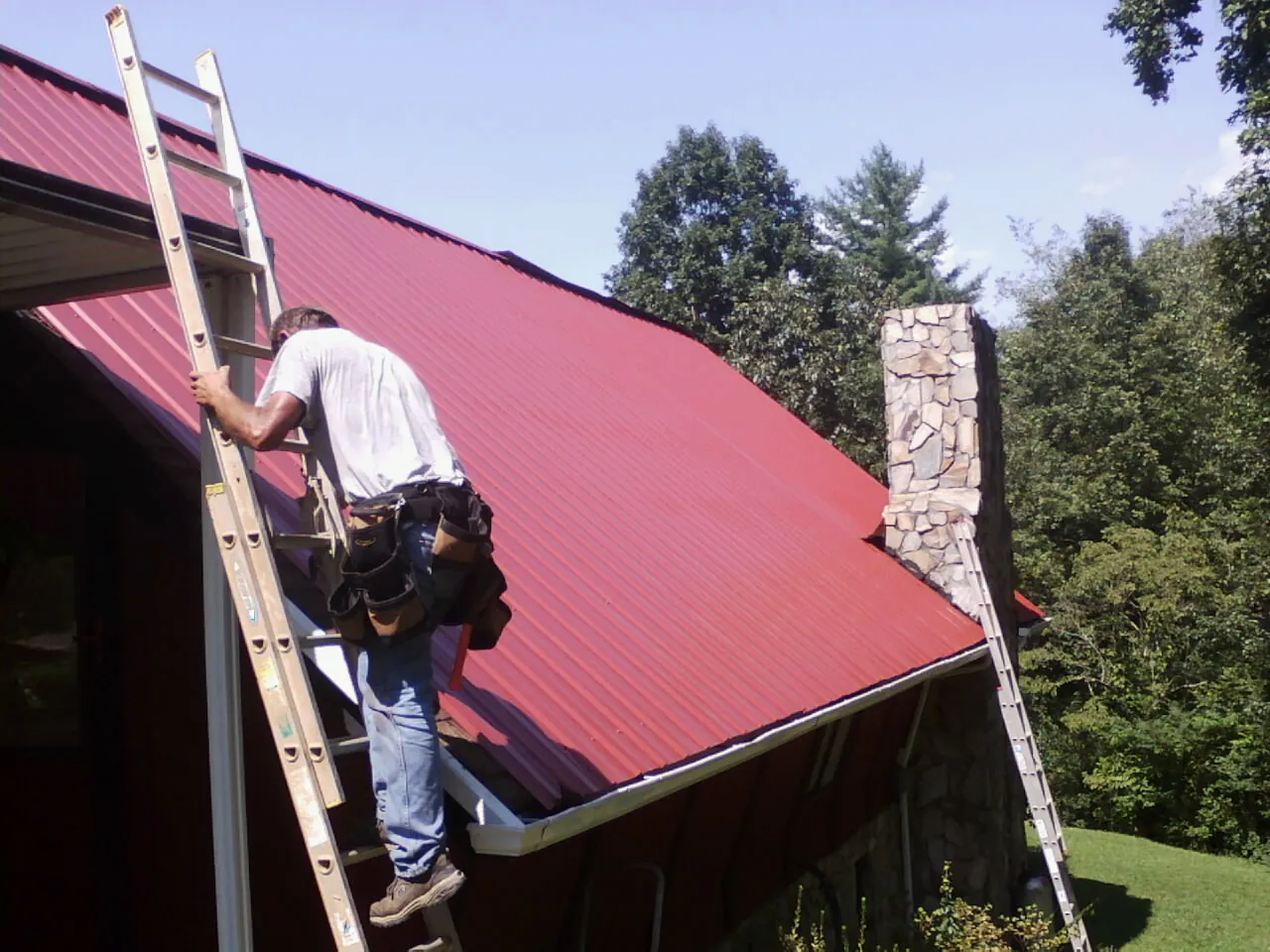 Roofer on a ladder beside a red metal roof and stone chimney — Soffit & Fascia Repair by Roofing Brothers in Mokena